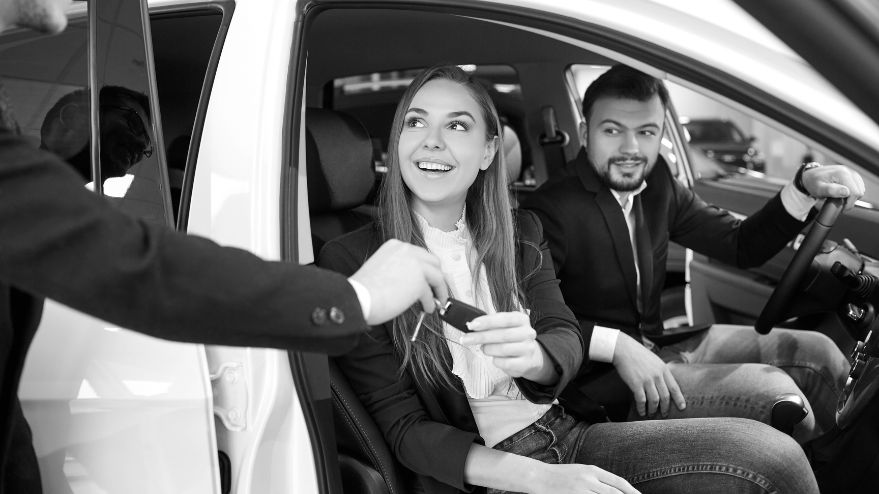 Young couple sitting in the front seats of a car inside a showroom, receiving the keys to their new vehicle from a salesperson.